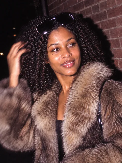 Fictional woman laughing beside a brick wall in a fur-collared coat, photographed with 1980s paparazzi flash